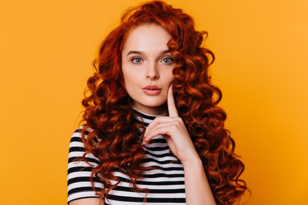 Girl with red curls and blue eyes touches her cheek with her finger and looks thoughtfully at camera on isolated orange background.